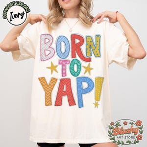 Puede incluir: Camiseta de color marfil con el texto "BORN TO YAP!" en letras coloridas y estampadas. La camiseta presenta estrellas y es de Blushy Art Store.