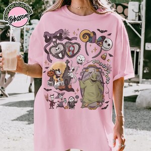 May include: Pink t-shirt featuring a colorful graphic design with characters and elements from "The Nightmare Before Christmas." The design includes Jack Skellington, Sally, Oogie Boogie, and other iconic imagery. The shirt has a relaxed fit.