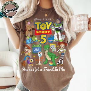May include: A brown short-sleeve t-shirt featuring a graphic print of Disney Pixar Toy Story characters including Woody, Buzz Lightyear, Jessie, Rex, and Forky. The text reads Toy Story 5 and You've Got a Friend In Me. A Comfort Colors Espresso logo is visible.