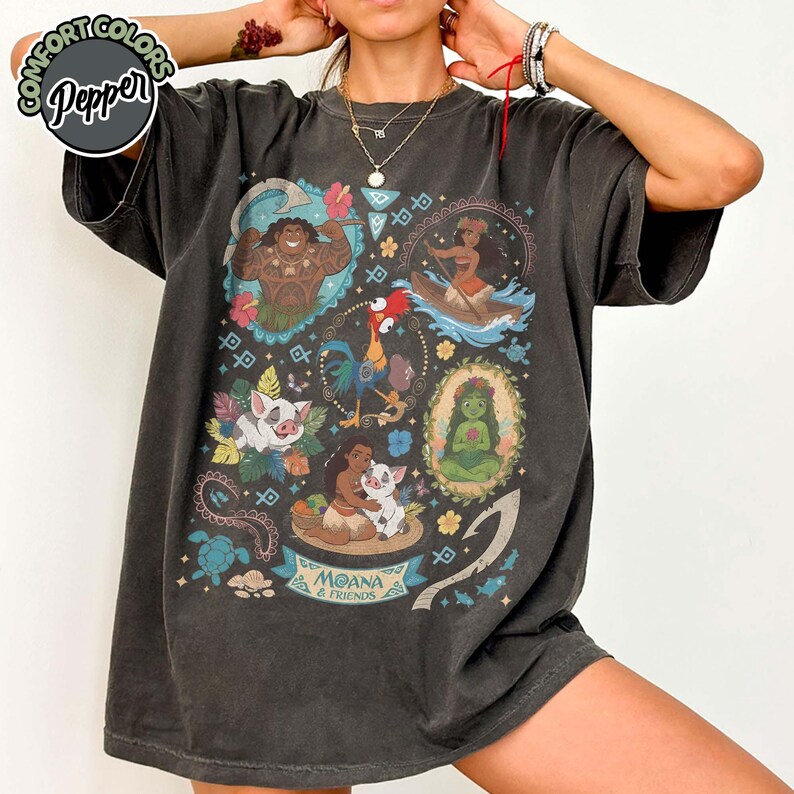May include: A charcoal gray short-sleeve t-shirt featuring a graphic print of Moana and friends, including Maui, Pua, Heihei, and Te Fiti. The design includes tropical flowers, tribal patterns, and a banner with the text Moana & Friends.