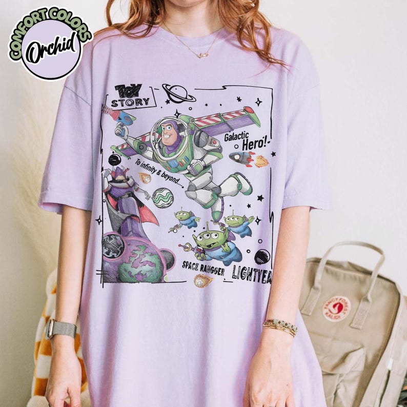Puede incluir: Una camiseta de color morado claro con un estampado gr&aacute;fico de Buzz Lightyear de Toy Story, junto con el texto "Toy Story", "Galactic Hero!", "To infinity & beyond....", "Space Ranger" y "Lightyear" rodeado de ilustraciones espaciales.