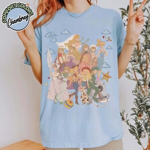 May include: A light blue crew neck t-shirt featuring a large graphic print of multiple animated female characters in various outfits, including a sheriff star, a rocket, and clouds. The shirt has a Comfort Colors Chambray label in the corner.