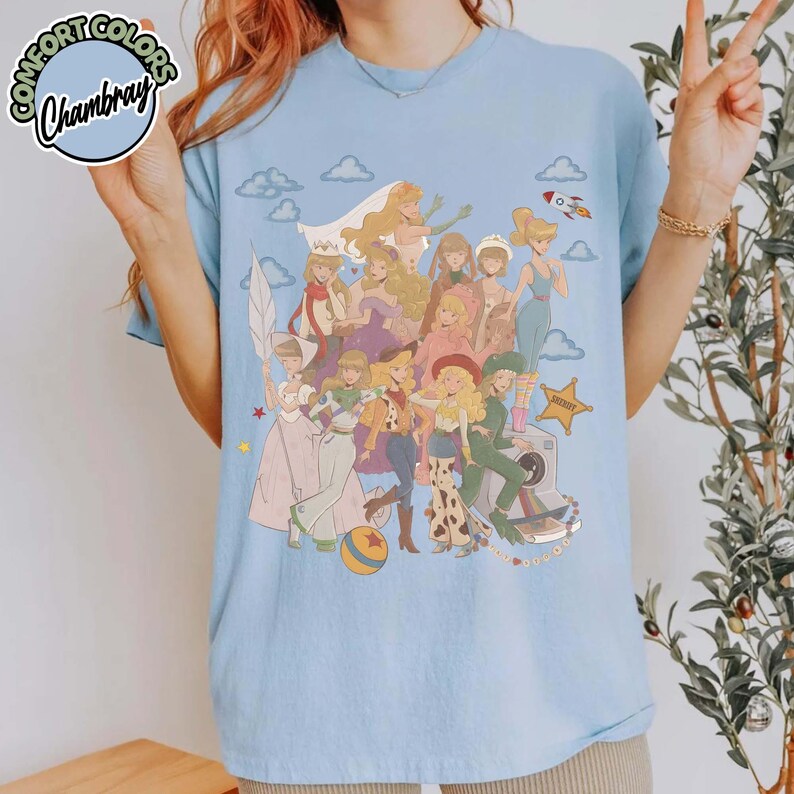 May include: A light blue crew neck t-shirt featuring a large graphic print of multiple animated female characters in various outfits, including a sheriff star, a rocket, and clouds. The shirt has a Comfort Colors Chambray label in the corner.