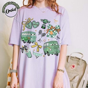 Puede incluir: Una camiseta de color lila claro con un dise&ntilde;o de dibujos animados de veh&iacute;culos, lazos y la palabra "Cars". El dise&ntilde;o incluye una furgoneta VW verde, una bandera a cuadros y un trofeo. La camiseta tiene las palabras "Comfort Colors" y "Orchid" en un c&iacute;rculo.