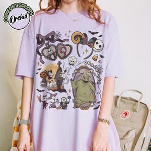 May include: A light purple t-shirt featuring a graphic design with characters and elements from "The Nightmare Before Christmas." The design includes Jack Skellington, Sally, Oogie Boogie, and other spooky elements.