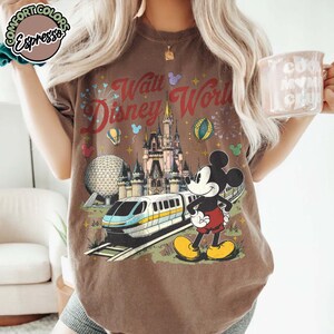May include: Brown t-shirt with a graphic of Mickey Mouse, Cinderella's castle, and a monorail, with the text "Walt Disney World". The design includes fireworks, balloons, and the Epcot ball. The shirt has the text "Comfort Colors" on the top left.