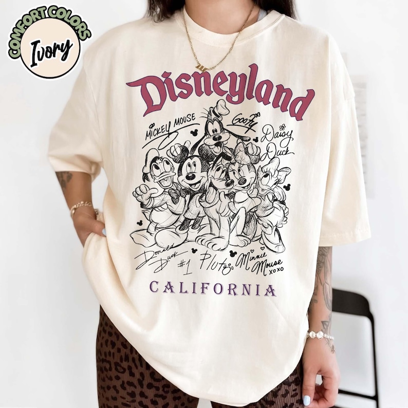 Puede incluir: Camiseta color marfil con un gr&aacute;fico de Disneyland. El dise&ntilde;o incluye a Mickey Mouse, Goofy, Donald Duck, Daisy Duck, Pluto y Minnie Mouse. Tambi&eacute;n se ve el texto "Disneyland" y "California".