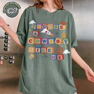 May include: A moss green t-shirt featuring a graphic of colorful alphabet blocks spelling out the text "YOU'RE A COWBOY LIKE ME". The design includes illustrations of a rocket, a sheriff star, a cowboy hat, and cowboy boots among small stars.
