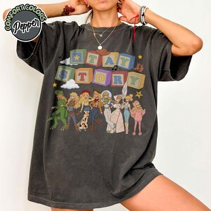May include: A charcoal grey short-sleeve t-shirt featuring the text STAY STORY in colourful block letters. The graphic includes illustrations of various characters dressed in costumes, along with small clouds, stars, and a musical note icon.