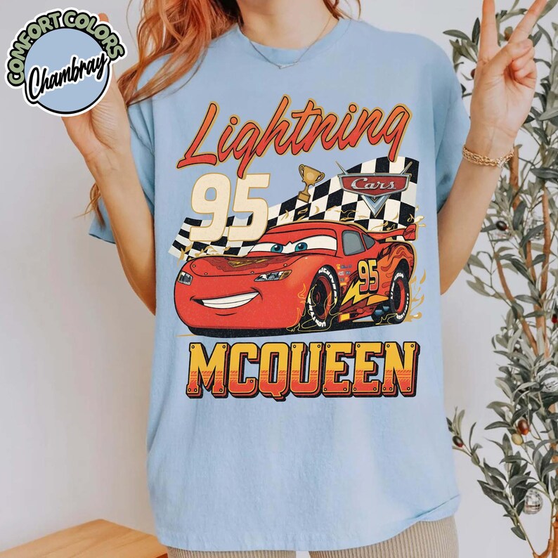 Pu&ograve; includere: T-shirt azzurra con una grafica di Saetta McQueen del film Cars. Il design include il testo "Lightning 95 McQueen" in arancione e giallo, con una bandiera a scacchi e un trofeo.