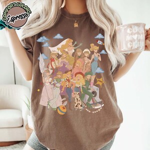 May include: A dark brown short-sleeve t-shirt featuring a large graphic print of multiple animated characters in various outfits, including a space suit, a cowgirl costume, and a ballgown, alongside a small rocket and a yellow star.