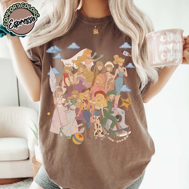 May include: A dark brown short-sleeve t-shirt featuring a large graphic print of multiple animated characters in various outfits, including a space suit, a cowgirl costume, and a ballgown, alongside a small rocket and a yellow star.