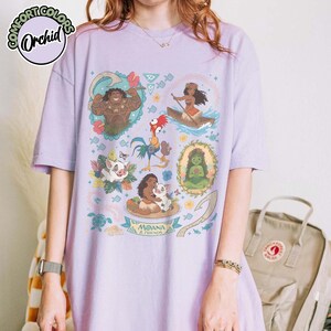 May include: A light purple short-sleeve t-shirt featuring a colorful graphic print of characters from Moana, including Maui, Moana, Pua, and Heihei. The design includes floral elements, a hook, and the text Moana & Friends in a central banner.