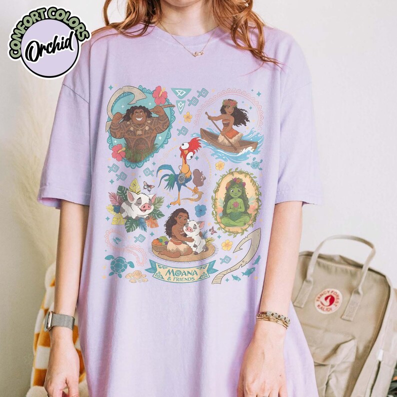May include: A light purple short-sleeve t-shirt featuring a colorful graphic print of characters from Moana, including Maui, Moana, Pua, and Heihei. The design includes floral elements, a hook, and the text Moana & Friends in a central banner.
