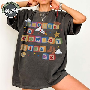 May include: A charcoal gray short-sleeve t-shirt featuring a graphic of colorful alphabet blocks spelling out the text YOU'RE A COWBOY LIKE ME. The design includes illustrations of a rocket, a sheriff star, a cowboy hat, and brown cowboy boots.