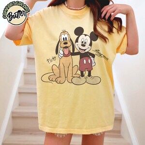 May include: Yellow t-shirt featuring a graphic of Mickey Mouse and Pluto. Mickey Mouse is wearing red shorts and Pluto is a tan color. The text "Mickey Mouse" is written on the shirt. The shirt has the text "Comfort Colors Butter" on the top left.