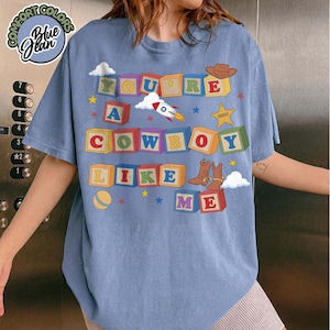 May include: A blue t-shirt featuring a graphic of colorful alphabet blocks spelling out the text YOU'RE A COWBOY LIKE ME. The design includes illustrations of a rocket ship, a sheriff star, a cowboy hat, cowboy boots, and a yellow ball.