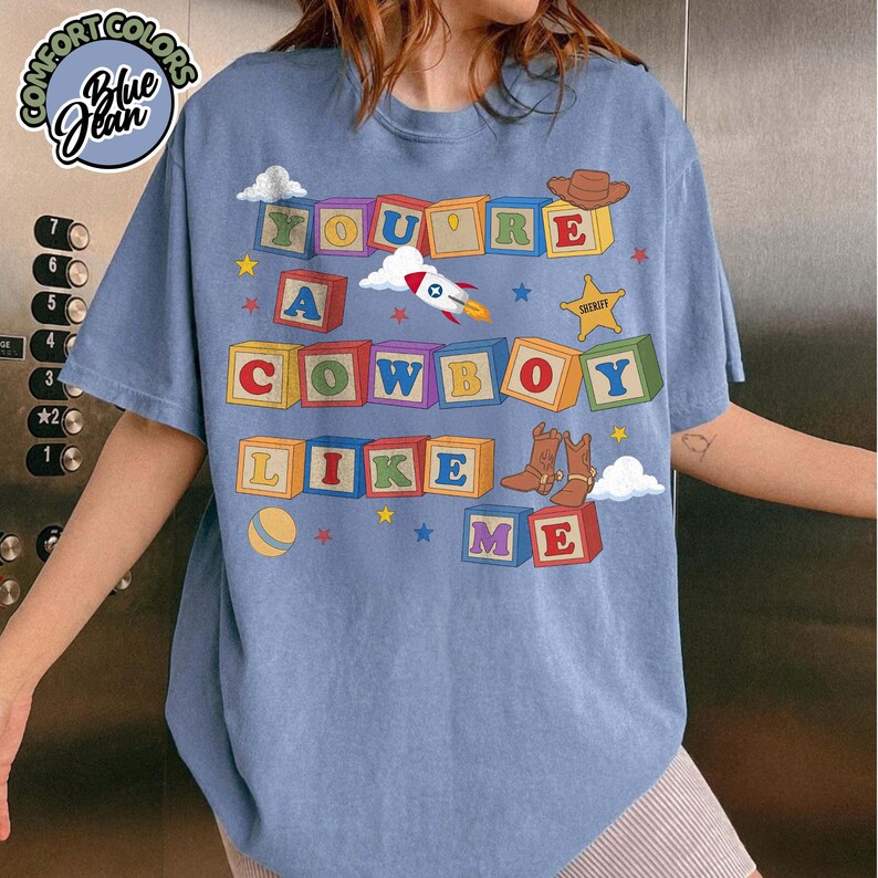 May include: A blue t-shirt featuring a graphic of colorful alphabet blocks spelling out the text YOU'RE A COWBOY LIKE ME. The design includes illustrations of a rocket ship, a sheriff star, a cowboy hat, cowboy boots, and a yellow ball.