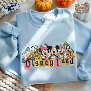 May include: A light blue crewneck sweatshirt featuring an embroidered graphic of Disney characters including Donald Duck, Pluto, Minnie Mouse, Mickey Mouse, Daisy Duck, and Goofy. The text Disney Land is embroidered below the characters in colorful blocks.