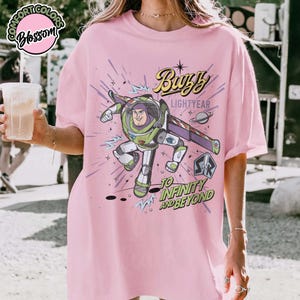 May include: Pink t-shirt featuring a graphic of Buzz Lightyear from Toy Story. The design includes the text "Buzz Lightyear" and "To Infinity and Beyond" with a space-themed background. The shirt has short sleeves and a relaxed fit.