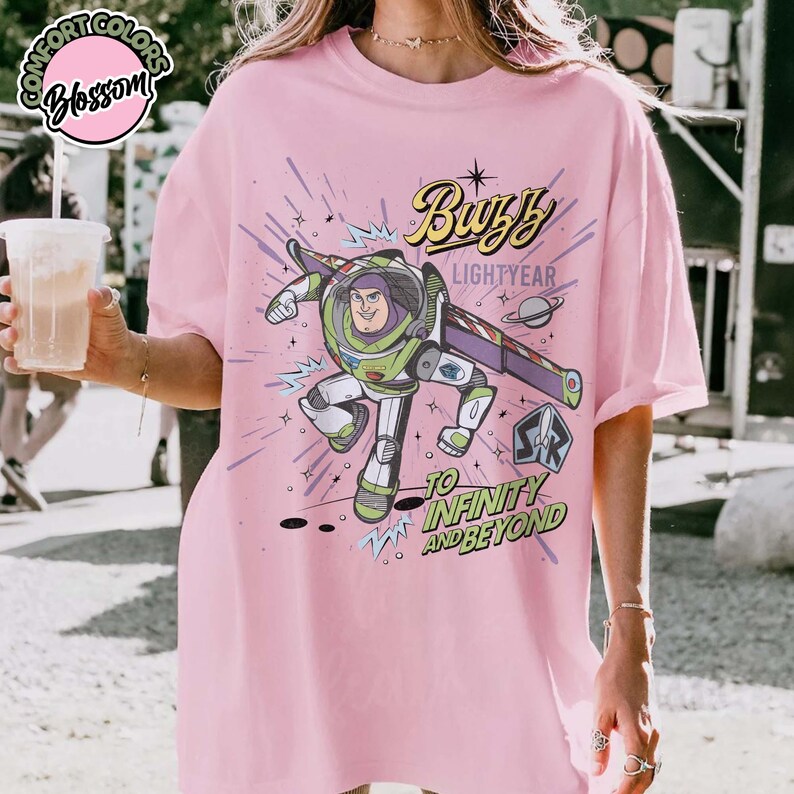 May include: Pink t-shirt featuring a graphic of Buzz Lightyear from Toy Story. The design includes the text "Buzz Lightyear" and "To Infinity and Beyond" with a space-themed background. The shirt has short sleeves and a relaxed fit.
