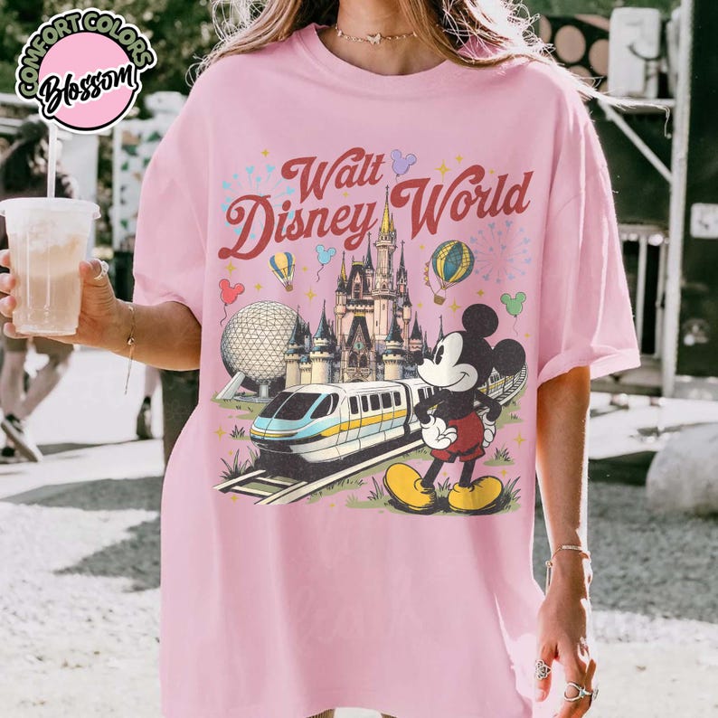 May include: Pale pink t-shirt with a vintage-style graphic of Walt Disney World, including Mickey Mouse, a monorail, and the castle. The text "Walt Disney World" is in a retro font. The shirt has a loose fit.