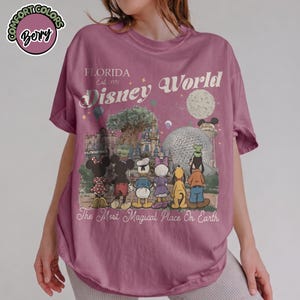 May include: A mauve-colored t-shirt featuring a graphic of Disney World characters and landmarks. The text "FLORIDA Est. 1971 Disney World" is printed above the graphic. The phrase "The Most Magical Place On Earth" is printed below the graphic.