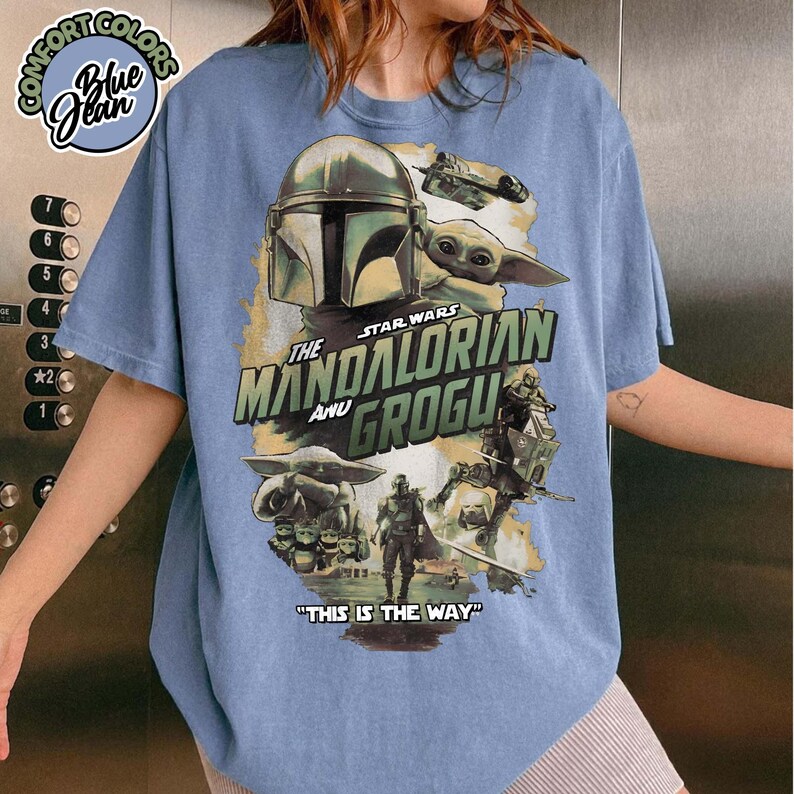 May include: A blue t-shirt featuring a graphic print of The Mandalorian and Grogu. The design includes the text "The Mandalorian and Grogu" and "This is the Way" along with illustrations of the characters, a spaceship, and a speeder bike.