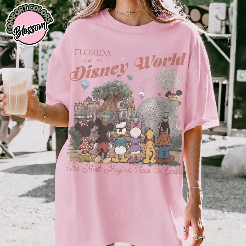 May include: Pink t-shirt with a graphic of Disney World characters and landmarks. The text "FLORIDA Est. 1971 Disney World" is printed above the graphic. The phrase "The Most Magical Place On Earth" is printed below the graphic.