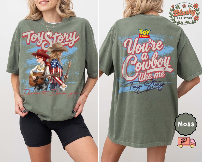May include: A moss green t-shirt featuring a Toy Story graphic. The front displays two cowgirls with guitars and the text I know it, I know you. The back shows the Toy Story logo with the text You're a Cowboy like me and Toy Story in script.