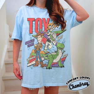 Puede incluir: Camiseta azul claro con un dise&ntilde;o de Toy Story que presenta a Woody, Buzz Lightyear y Rex. El dise&ntilde;o incluye el texto "TOY STORY" en rojo, "SINCE 1995" y los nombres de los personajes. La camiseta tiene un corte holgado.