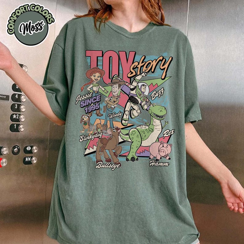 May include: Moss green t-shirt featuring a graphic of Toy Story characters, including Woody, Buzz, and Rex. The design includes the text "TOY STORY" and "Jessie SINCE 1995". The shirt has a relaxed fit.