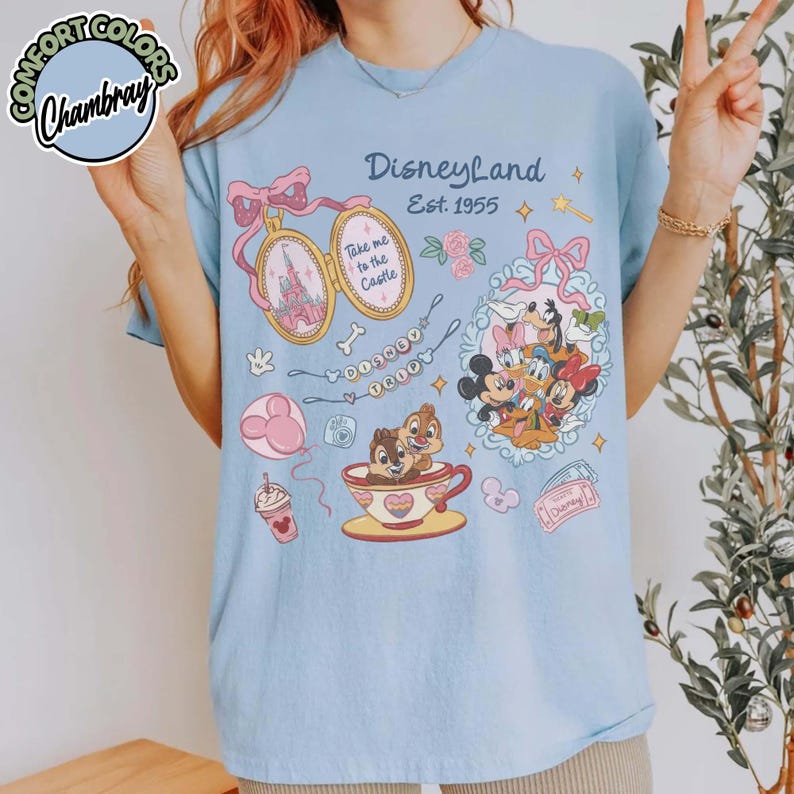Peut inclure: T-shirt bleu clair avec un motif Disneyland. Le motif comprend un ch&acirc;teau, Mickey Mouse, Minnie Mouse, Donald Duck et Dingo. Le texte "Disneyland Est. 1955" est &eacute;galement visible.