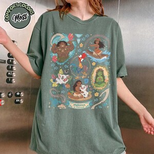 May include: A moss green short-sleeve t-shirt featuring a graphic print of Moana and Friends characters including Maui, Pua, Heihei, and Te Fiti. The design includes floral accents, tribal patterns, and the text Moana & Friends.