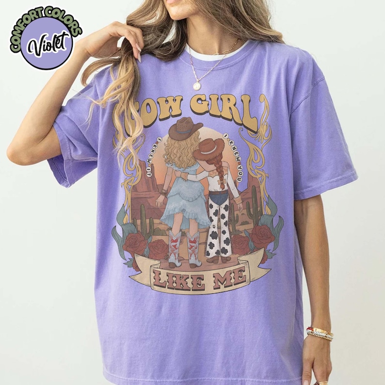 May include: A violet short-sleeve t-shirt featuring a graphic print of two figures in cowboy hats and boots standing in a desert landscape with cacti and roses. The text reads COW GIRL LIKE ME and I KNEW YOU. A circular logo in the corner says COMFORT COLORS Violet.