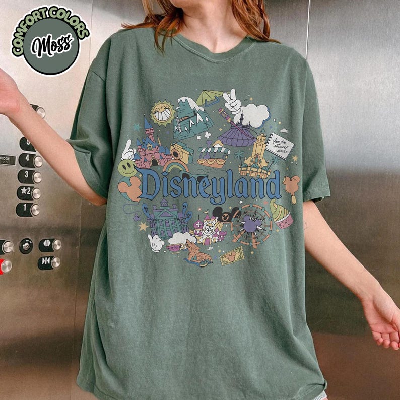 Puede incluir: Camiseta verde musgo con un gr&aacute;fico de Disneyland. El gr&aacute;fico presenta varios elementos de dibujos animados, incluyendo un castillo, un sol y la palabra "Disneyland". La camiseta tiene mangas cortas y un corte holgado.