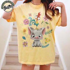 May include: Yellow t-shirt featuring a cartoon pig and rooster with floral accents. The shirt has the text "Comfort Colors Butter" in a circular design at the top left. The design includes pink, yellow, and blue flowers.
