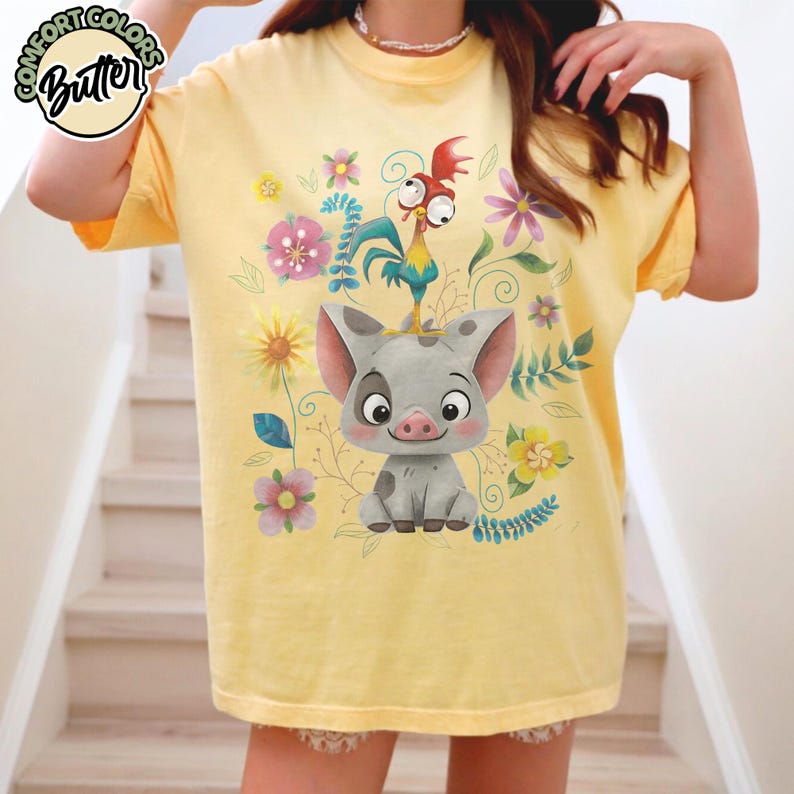 May include: Yellow t-shirt featuring a cartoon pig and rooster with floral accents. The shirt has the text "Comfort Colors Butter" in a circular design at the top left. The design includes pink, yellow, and blue flowers.