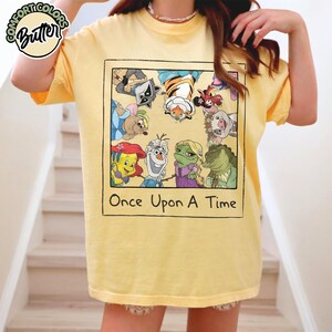 Puede incluir: Camiseta amarilla con un gr&aacute;fico que presenta personajes de dibujos animados dentro de un dise&ntilde;o enmarcado. El texto "Once Upon A Time" est&aacute; debajo de la imagen. La camiseta tiene mangas cortas y cuello redondo.
