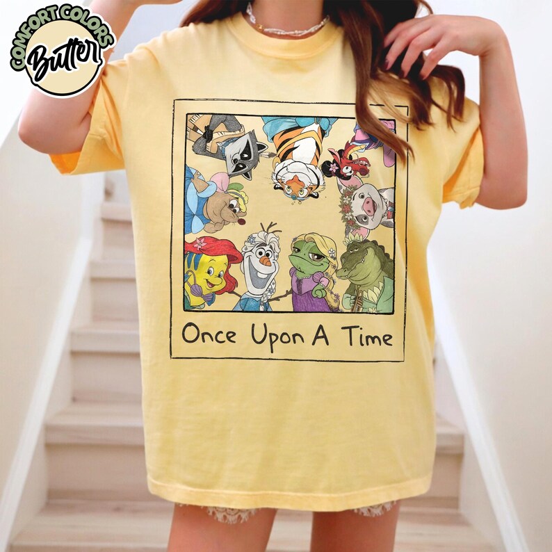 Puede incluir: Camiseta amarilla con un gr&aacute;fico que presenta personajes de dibujos animados dentro de un dise&ntilde;o enmarcado. El texto "Once Upon A Time" est&aacute; debajo de la imagen. La camiseta tiene mangas cortas y cuello redondo.