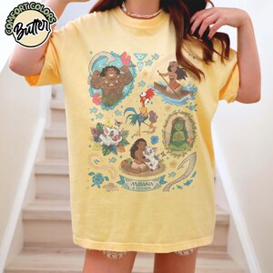 May include: A pale yellow t-shirt featuring a graphic print of characters from the movie Moana, including Maui, Moana, Hei Hei, Pua, and Te Fiti. The design includes tropical flowers, a turtle, and a fishhook, with the text Moana & Friends.