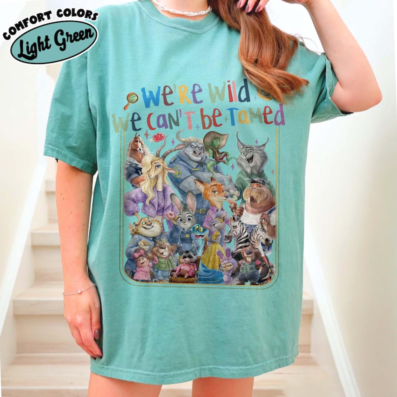 May include: Light green t-shirt with the text "We're Wild, We Can't Be Tamed" and a colorful graphic of animated animal characters. The shirt is a comfort color and has a relaxed fit.