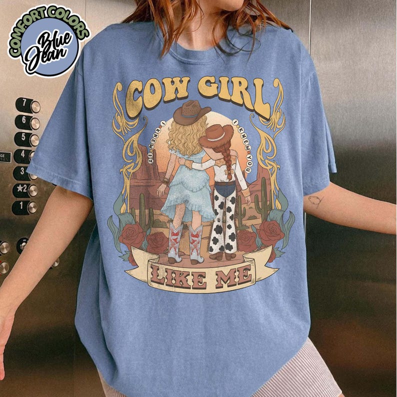 May include: A blue short-sleeve t-shirt featuring a graphic of two figures in cowboy hats standing in a desert landscape with cacti and roses. The text reads COW GIRL LIKE ME and I KNEW IT I KNEW YOU. A Comfort Colors Blue Jean logo is in the corner.