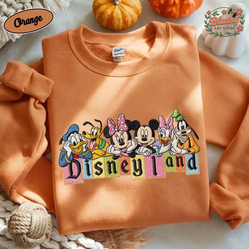 May include: An orange crewneck sweatshirt featuring an embroidered graphic of Disney characters including Donald Duck, Pluto, Minnie Mouse, Mickey Mouse, Daisy Duck, and Goofy. Below the characters, the text Disneyland is embroidered in black.