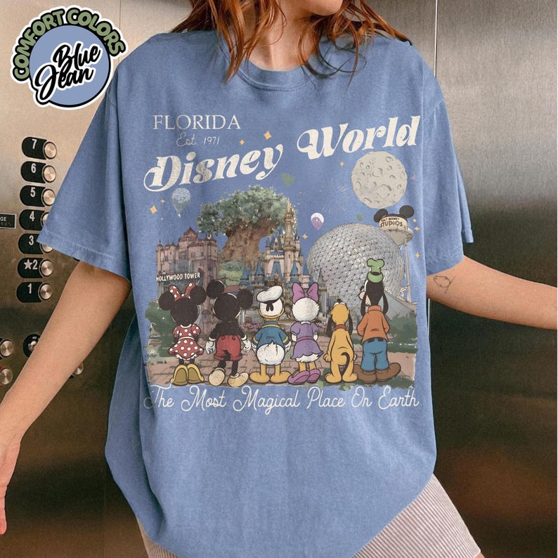 May include: Blue short-sleeved t-shirt with a graphic of Disney World in Florida. The design includes the text "Florida Est. 1971 Disney World" and features cartoon characters and park landmarks. The shirt also has the text "The Most Magical Place On Earth."