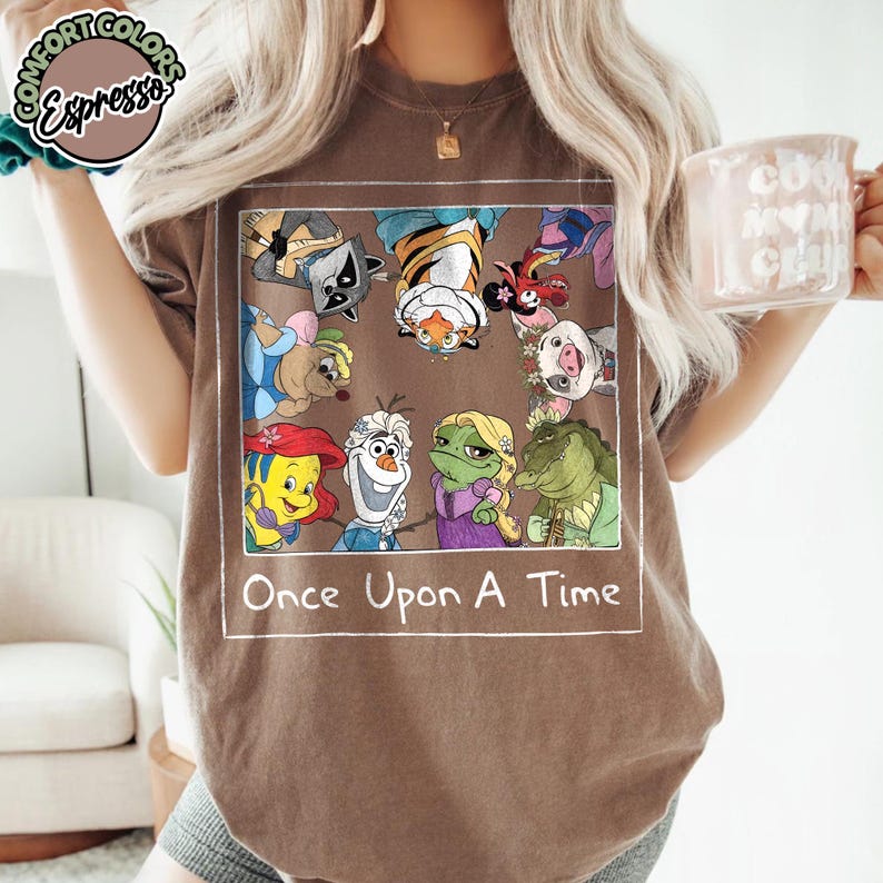Puede incluir: Una camiseta marr&oacute;n con un gr&aacute;fico de personajes de dibujos animados. El gr&aacute;fico est&aacute; enmarcado en un rect&aacute;ngulo blanco e incluye el texto "Once Upon A Time". La camiseta es de la marca Comfort Colors.