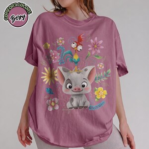 May include: Mauve t-shirt featuring a graphic of a cartoon pig, rooster, and flowers. The pig is gray with dark spots and large ears. The rooster is blue, yellow, and red. The shirt has the text "Comfort Colors" and "Bory" in a circle.