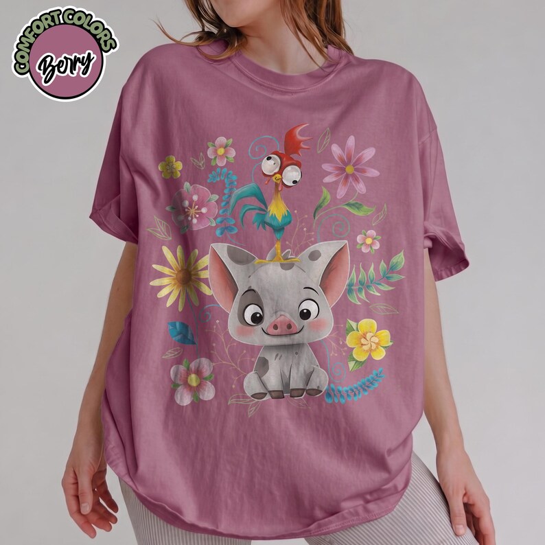 May include: Mauve t-shirt featuring a graphic of a cartoon pig, rooster, and flowers. The pig is gray with dark spots and large ears. The rooster is blue, yellow, and red. The shirt has the text "Comfort Colors" and "Bory" in a circle.