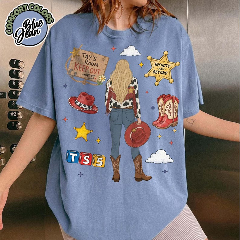 May include: A blue short-sleeve t-shirt featuring a graphic of a person with long blonde hair wearing a cow-print jacket and jeans. The design includes a sign reading TAY'S ROOM KEEP OUT, a yellow star badge, red cowboy boots, and blocks labelled TS5.