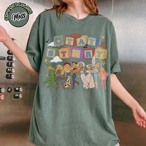 May include: A moss green short-sleeve t-shirt featuring a graphic print of colourful alphabet blocks spelling out "TAY STORY" above a line of illustrated characters dressed in various outfits, including a dinosaur suit and space-themed attire.